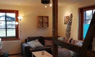 A cozy living room with a large sofa, a wooden table, and a lamp. Bright walls and windows create a friendly atmosphere.
