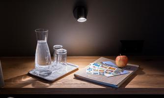 A table with a glass carafe, two glasses, and a red apple. On top of it lies a notebook with postcards.