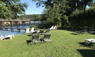 A green meadow with deck chairs and a view of a calm lake. In the background, there are trees and small wooden houses.