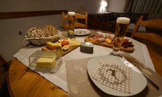 A cozy table with various types of cheese, sausage, and fresh bread. Two glasses of beer and a plate are ready on the side.