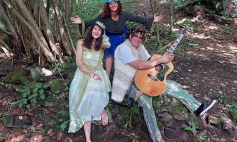 Drei Personen sitzen im Wald auf einem Stein. Eine Frau trägt ein festliches Kleid, während ein Mann ein akustisches Gitarrenspiel macht.