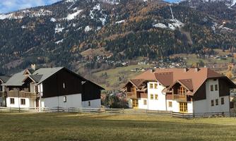 A picturesque landscape with mountains in the background and modern, traditional houses. The meadow is green and the trees display autumn colors.