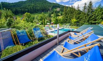 A beautiful terrace with loungers and a view of the green mountains. In the background, trees and a swimming pool are visible.