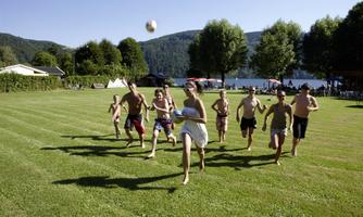 A group of young people is playing outdoors with a ball. The scene shows a green meadow and a clear lake in the background.