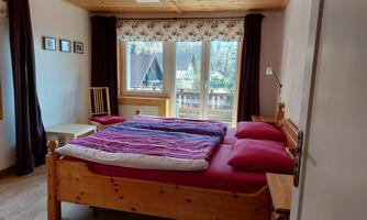 A cozy bedroom with a double bed and red bed linen. Large windows offer a view of the outdoors and let in plenty of daylight.