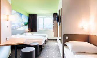 A modern hotel room with two beds and an additional sleeping space. Large windows let in plenty of light, and the decor is simple and functional.