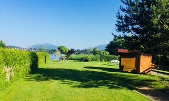 A beautiful garden with green grass, trees, and a view of the lake. In the background, mountains are visible, and the sun is shining in the clear sky.