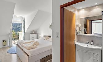 A bright bedroom with a large bed and access to a balcony. In the background, you can see a modern bathroom with a sink and a mirror.