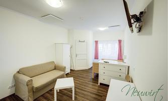 A bright room with a beige sofa, a table, and a TV on the wall. The room has wooden flooring and large windows with red curtains.