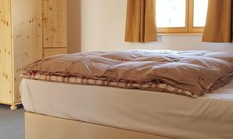 A simple bedroom with a bed, a wooden wardrobe, and a window with light curtains. The room is well-lit and tidy.