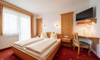 A cozy hotel room with two single beds, a desk, and a television. Large windows provide plenty of daylight.