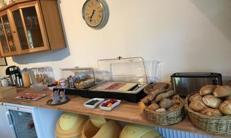 A colorful breakfast buffet with various rolls, cold cuts, and fresh fruit. In the background, there is a kitchen clock and a beverage dispenser.