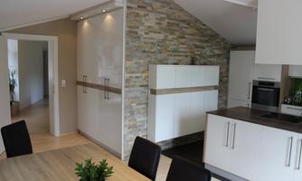 Modern kitchen with elegant white furniture and a stone wall accent. A dining area with a wooden table and chairs is also visible.