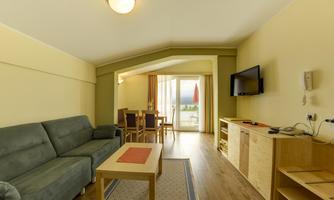 A bright living room with a gray sofa and a table. The large windows provide access to a balcony with a view of the outdoors.