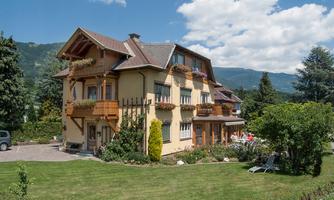 A beautiful two-story house with a balcony and a green garden. In the background, there are mountains and a blue sky.