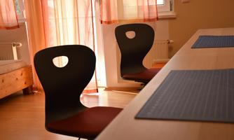 A bright room with two ergonomic chairs and a table. The windows are decorated with orange curtains.
