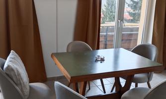 A cozy dining area with a wooden table and upholstered chairs. A television hangs on the wall and large windows offer a view of the outdoor area.