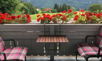 A balcony with two chairs and a small table, surrounded by red flowers. In the background, green meadows and mountains are visible.