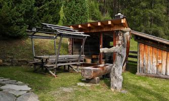 A rustic wooden cabin with outdoor seating. Surrounded by trees and a green meadow.