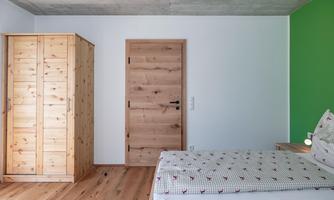 A modern bedroom with wooden furniture and a green accent wall. A single bed and a wardrobe are visible in the room.