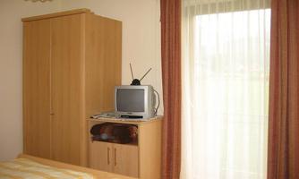 A simple bedroom with two beds, a wooden wardrobe, and a small television. Daylight enters the room through a window with curtains.