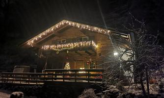A cozy wooden house at night, adorned with lights. The snow covers the ground and the surroundings, creating a festive atmosphere.
