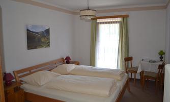 A cozy bedroom with two single beds and a table in the corner. Large windows allow plenty of daylight in.