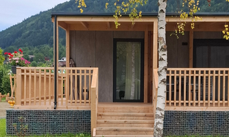 A modern wooden house with a wooden porch and a view of the surrounding mountains. In front of the house are some flowers and trees.