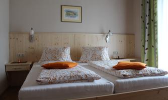 A cozy bedroom with two single beds and orange pillows. The walls are light, and the wooden furniture creates a warm atmosphere.