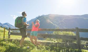 A couple stands by a wooden fence in the mountains. They enjoy the sun and the beautiful scenery.