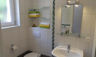 A modern bathroom with white tiles and a sink. Towels in various colors are neatly arranged on a shelf.