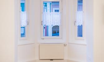 A bright room with three windows and white curtains. The floor is made of wood and there is a radiator under the central window.