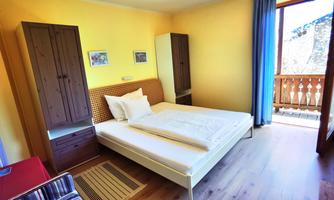 A cozy bedroom with a double bed and a large window. The walls are painted yellow, and there are wooden cabinets as well as a balcony with a view outside.