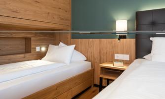 A modern bedroom with two single beds and wooden decor. There is a bedside lamp and a small table next to one bed.