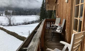 A balcony with two garden chairs and a view of a snow-covered landscape. In the background, trees and mountains can be seen, while the sky is cloudy.