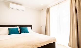 A modern bedroom with a large bed and pillows in blue. Large windows with curtains let in plenty of daylight.