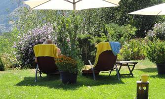 Two lounge chairs are located in the garden under a sun umbrella. Surrounded by colorful flowers and green grass, the scene invites relaxation.