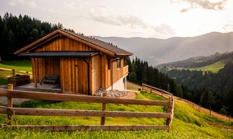 A cozy wooden house on a meadow with a wide view of the mountains. The landscape is green and accompanied by a dramatic sky.