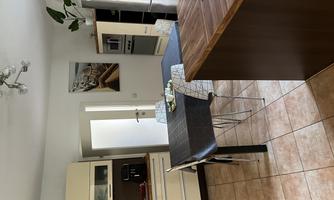 A modern kitchen with a dining table and simple furniture. The walls are light, and there are windows that let in plenty of light.