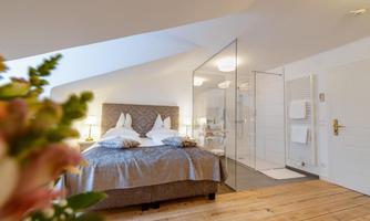 A modern bedroom with a large bed and soft lighting. Next to the bed, there is a transparent shower bath.