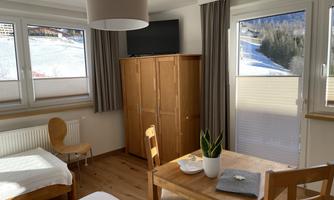 A cozy room with wooden furniture and large windows. The view shows snow and trees in the background.