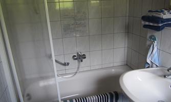 A bathroom with a bathtub and a shower. Next to the sink, towels are on a shelf.