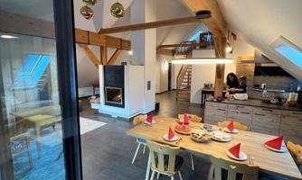 A cozy dining area with a wooden table and chairs. On the table, there are Christmas hats and some food, while a modern kitchen can be seen in the background.