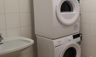 A washing machine and a dryer are stacked on top of each other in a bright room. Next to them is a sink.