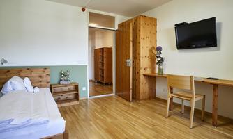A cozy room with wooden furniture and a large window. The wall is painted in soft colors and a television is mounted.