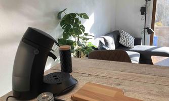 A modern kitchen corner with a coffee machine, cutting board, and decorative candles. In the background, there is a cozy living area with a gray couch and plants.