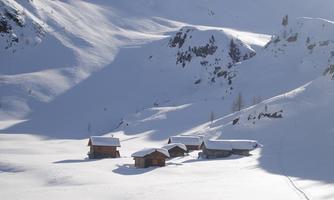 A tranquil winter landscape with several cabins, surrounded by fresh snow. The mountains in the background are visible with snow-covered peaks.