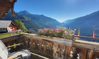 A beautiful view from a balcony overlooking a valley with mountains in the background. Colorful flowers and a cozy seating area complete the picture.