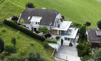 A modern house with a well-maintained garden and many plants. In the background, a green meadow can be seen.