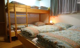 A cozy bedroom with a bunk bed and two single beds. The bedding is colorful and the windows let in soft light.